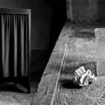 Photographie épurée en noir et blanc présentant un isoloir au rideau tiré et, séparés par une poutre en bois au sol, une urne transparente sans inscription avec un bulletin de vote froissé abandonné par terre.