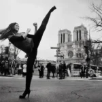Une photographie en noir et blanc montre une femme effectuant un coup de pied haut et puissant sur une rue de Paris. Ses longs cheveux volent en arrière et elle sourit, portant des leggings noirs et un débardeur texturé. Sa jambe droite est levée très haut, presque à la verticale. Elle se tient sur son pied gauche qui porte un talon aiguille. En arrière-plan, la cathédrale Notre-Dame de Paris est visible avec ses deux tours. Des passants, un bus OpenTour à impériale et un grand arbre nu complètent la scène urbaine. Le sol de la rue est usé et texturé.