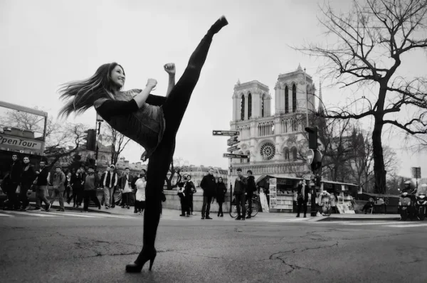 Une photographie en noir et blanc montre une femme effectuant un coup de pied haut et puissant sur une rue de Paris. Ses longs cheveux volent en arrière et elle sourit, portant des leggings noirs et un débardeur texturé. Sa jambe droite est levée très haut, presque à la verticale. Elle se tient sur son pied gauche qui porte un talon aiguille. En arrière-plan, la cathédrale Notre-Dame de Paris est visible avec ses deux tours. Des passants, un bus OpenTour à impériale et un grand arbre nu complètent la scène urbaine. Le sol de la rue est usé et texturé.