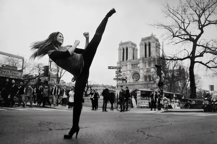 Une photographie en noir et blanc montre une femme effectuant un coup de pied haut et puissant sur une rue de Paris. Ses longs cheveux volent en arrière et elle sourit, portant des leggings noirs et un débardeur texturé. Sa jambe droite est levée très haut, presque à la verticale. Elle se tient sur son pied gauche qui porte un talon aiguille. En arrière-plan, la cathédrale Notre-Dame de Paris est visible avec ses deux tours. Des passants, un bus OpenTour à impériale et un grand arbre nu complètent la scène urbaine. Le sol de la rue est usé et texturé.
