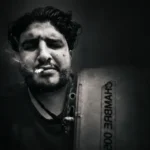 Portrait sombre en noir et blanc d'un homme une cigarette aux lèvres, tenant un panneau où est écrit « CHAMBRE 002 », dans une ambiance dark et contrastée.