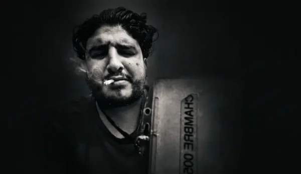 Portrait sombre en noir et blanc d'un homme une cigarette aux lèvres, tenant un panneau où est écrit « CHAMBRE 002 », dans une ambiance dark et contrastée.