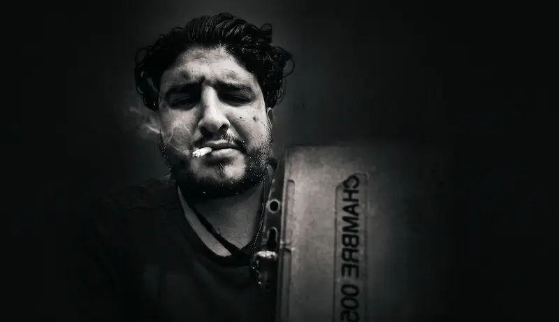 Portrait sombre en noir et blanc d'un homme une cigarette aux lèvres, tenant un panneau où est écrit « CHAMBRE 002 », dans une ambiance dark et contrastée.